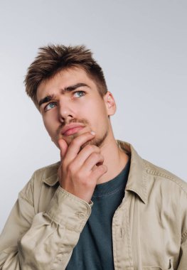 Kısa saçlı genç bir adam düşünceli bir şekilde poz veriyor, çenesini elinin üzerine koyuyor. Düşüncelere dalmış gibi görünüyor. Sıradan bir şekilde ceket ve süveter giymiş..