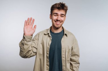 Açık kahverengi saçlı genç bir adam kapalı alanda kameraya gülümseyip el sallıyor. Günlük kıyafetleri arasında bej bir ceket ve yeşil bir gömlek var. Hoş bir ortam yaratıyor..