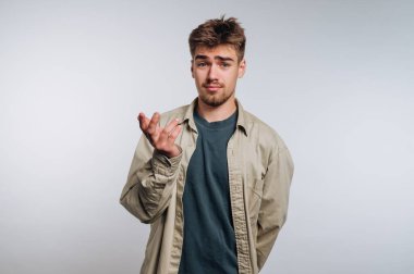 Genç bir adam sorgulama jesti için sol elini hafifçe kaldırdı. Şaşkın ya da kararsız görünüyor, düğmeli ceketin altına yeşil bir gömlek giyiyor. Arkaplan tarafsız.