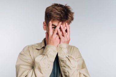 Genç bir adam iyi ışıklandırılmış bir stüdyoda duruyor, elleriyle yüzünü örtüyor. Açık renkli bir gömlek giyip utangaçlık ve utanç ifadesi sergiliyor..