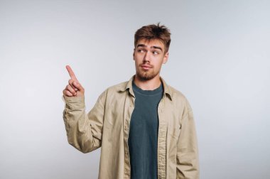 Genç bir adam açık gri bir stüdyoda duruyor, parmağını yukarı kaldırırken düşünceli görünüyor. Sıradan giyinmiş, rahat bir hava yayıyor..