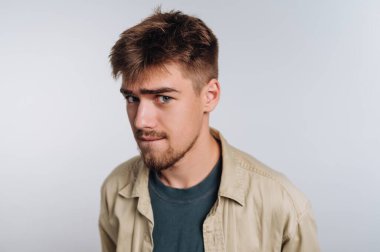 Genç bir adam kendine güvenerek duruyor, neşeli bir ifade sergiliyor hafif bir sırıtışla. Kısa, dağınık saçları ve sakalı var. Tişörtün üzerine sıradan bir tişört giymiş. Tarafsız bir arkaplanı var..