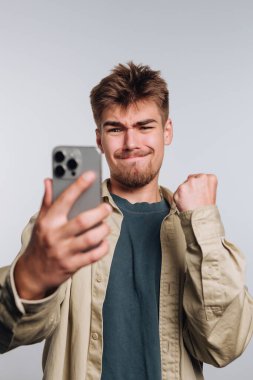 Parlak ve basit bir arka planda, genç bir adam selfie çekerken gülümser, bir yumruk pompasıyla mutluluğu ifade eder. Günlük kıyafetleri rahatlatıcı bir havayı yansıtıyor, bir neşe anı yakalıyor..