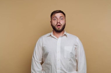 Sakallı bir adam beyaz gömlek giyen sade bir arka plana karşı duruyor. Gözleri kocaman ve ağzı açık, şaşırdığını ya da inanmadığını ifade ediyor..