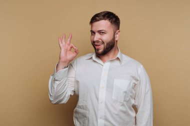 Sakallı bir adam beyaz gömlek giyerken özgüvenle gülümsüyor. Açık bej arkaplana karşı elini kullanarak onaylayıcı bir jest yapıyor. Atmosfer neşeli ve pozitif..