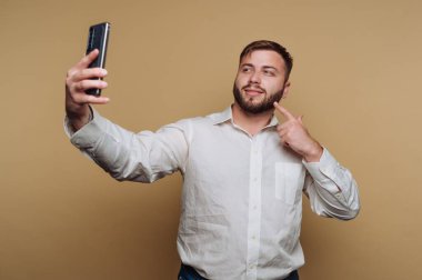 Genç bir adam akıllı telefonuyla selfie çekerken kendinden emin bir şekilde gülümsüyor. Şakacı bir şekilde poz veriyor, yüz ifadesini işaret ediyor. Arka plan katı, sıcak bir renktir..