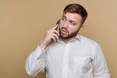 Beyaz gömlek giyen bir adam telefonda konuşuyor, düşünceli bir yüz ifadesi sergiliyor. Odaklanmış tavrı önemli bir konuşma yaptığını gösteriyor. Muhtemelen sıradan bir konuşma..