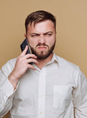 Sakallı bir adam telefonda konuşurken kaşlarını çatıyor. Beyaz bir gömlek giyiyor ve hayal kırıklığına uğramış görünüyor. Arka plan yumuşak, nötr bir renktir..