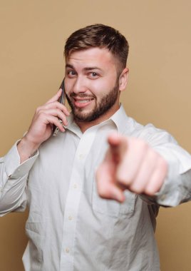 Sakallı genç bir adam telefonda konuşurken kendinden emin bir şekilde gülümsüyor. Direkt olarak kamerayı göstererek basit bir ortamda canlı ve ilgi çekici bir sahne yaratıyor..
