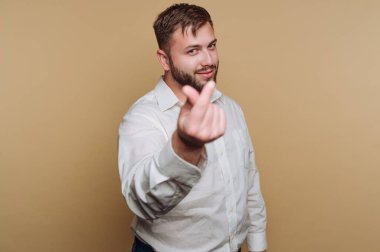 Genç bir adam sade bir arkaplanda duruyor, sıcaklığı ve dostluğu ifade eden bir el hareketi yapıyor. Günlük kıyafetleri ve gülüşü davetkar atmosfere katkı sağlıyor..