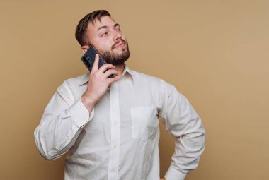 Beyaz gömlekli bir adam bir eli kalçasında, kulağına akıllı telefon dayamış duruyor. Düşünceli ve sohbete dalmış görünüyor. Rahat bir ortam yaratıyor..