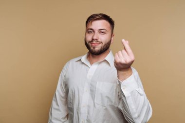 Neşeli bir adam açık kahverengi bir arka fona karşı duruyor. Beyaz düğmeli bir gömlek giyiyor ve şakacı bir el hareketi yapıyor, dostça bir hava veriyor..