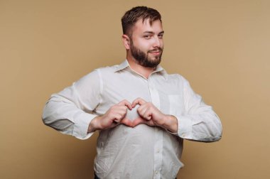 Bir adam gülümsüyor ve elleri önünde kalp şekli oluşturuyor. Beyaz düğmeli bir gömlek giyiyor ve tarafsız bir arkaplanda neşeli bir atmosfer yaratıyor..