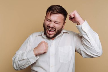 Bir adam kutlama için yumruklarını kaldırarak heyecan ve neşe sergiler. Onun geniş bir gülümsemesi ve anlamlı gözleri var. Arkaplan, onun coşkulu tavırlarını vurgulayan sade bir açık renk..