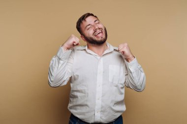 Sakallı bir adam gülümsüyor ve yumrukları havada tezahürat yapıyor. Açık renkli bir gömlek giyiyor ve açık bir arkaplanda duruyor, neşe ve heyecan taşıyor..