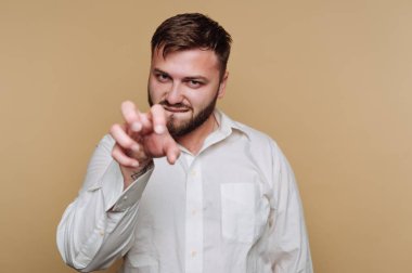 Sakallı bir adam kıskaç hareketi yapıyor, hafifçe gülümsüyor. Beyaz düğmeli bir gömlek giyiyor ve arka planı yumuşak bir bej rengi..