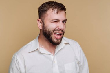 Sakallı bir adam göz kırpıyor ve gülümsüyor, neşeli bir kişilik sergiliyor. Beyaz bir gömlek giyiyor ve ışığın önünde duruyor. Tarafsız bir arkaplan..