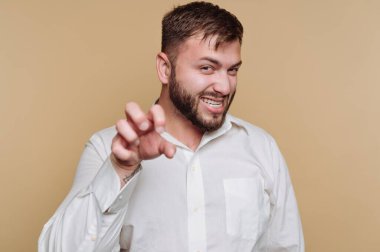 Sakallı ve kısa saçlı bir adam neşeli bir pençe hareketi yaparken geniş bir şekilde gülümsüyor. Beyaz bir gömlek giyer ve sade bir sarı arkaplan önünde poz verir, eğlenceli bir ruh yakalar..