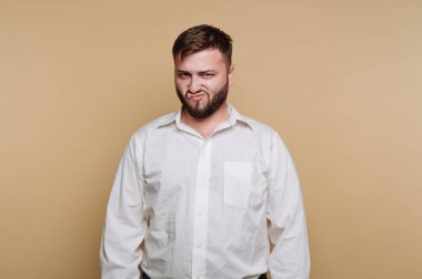 Bir adam basit bir geçmişi olan sıradan bir mekanda duruyor. Beyaz bir gömlek giyer ve şaşkın bir ifade sergiler, karışıklık ya da yaramazlık hissi taşır..