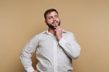 Genç bir adam tek eli çenesinde dalgın dalgın bakıyor. Beyaz bir gömlek giyiyor ve bej bir duvara karşı nötr bir ifade sergiliyor. Düşünceli duruşu iç gözlem olduğunu gösteriyor..