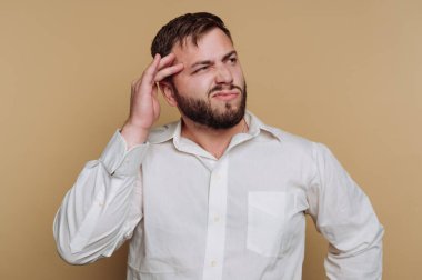 Kafasına dokunurken sakallı bir adamın kafası karışmış gibi görünüyor. Beyaz bir gömlek giyer ve bej bir arka plana yaslanır. Derin düşüncelere dalmayı ya da belirsizliği gösterir..