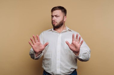 Sakallı bir adam iki elini de savunmacı bir şekilde kaldırıyor, bej bir arka fonun önünde duruyor. İfadesi bu durumda belirsizlik ya da rahatsızlık veriyor..