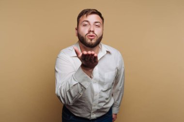 Sakallı bir adam açık renkli bir odada dururken şakacı bir şekilde öpücük yolluyor. Düğmeli bir gömlek giyiyor ve neşeli bir ifade sergiliyor, anı güzelleştiriyor..