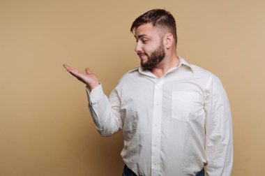 Bir adam sade bir arka planda güvenle durur. Beyaz bir gömlek giyer ve bir noktayı ifade etmek için eliyle işaret ederken soluna bakar. Davranışları rahat ve cana yakın..