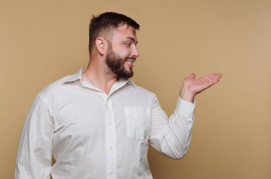 Sakallı bir adam bej bir arka fona karşı duruyor, gülümsüyor ve elini uzatıyor sanki bir şey gösteriyor gibi. Beyaz gömleği sıcak arka planla çelişiyor, sahneyi davetkar kılıyor..