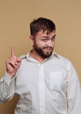 Sakallı bir adam bir parmağını şakacı bir şekilde kaldırır, kendine güvenerek gülümser. Beyaz bir gömlek giyiyor ve açık kahverengi bir arka plana karşı duruyor. Sıradan ve çekici bir atmosfer yaratıyor..