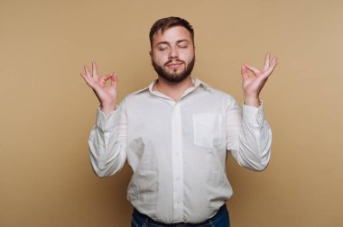 Sakallı bir adam huzurlu bir şekilde duruyor, gözleri kapalı ve elleri meditasyonla hareket ediyor. Düz zemin, onun meditasyona odaklanmasını artıracak sakin bir ortam yaratıyor..