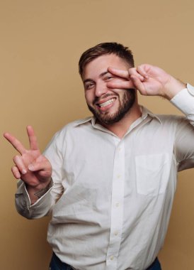 Neşeli bir adam bej bir arka fonun önünde duruyor, parmaklarıyla iki barış işareti yaparken geniş bir şekilde gülümsüyor. Kendinden emin ve rahat görünüyor. Olumlu bir hava yayıyor..