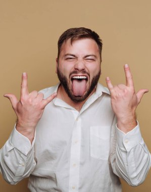 Beyaz gömlek giyen bir adam dilini uzatarak ve kaya işaretini elleriyle göstererek neşeli bir ifade takınır. Sakalı var ve mutlu poz veriyor..