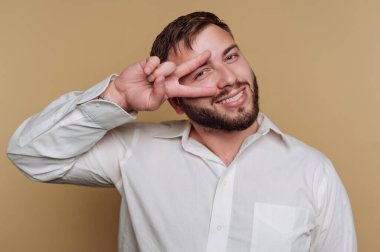 Beyaz gömlek giyen genç bir adam barış işareti ile poz verirken parlak bir şekilde gülümsüyor. Arka plan, onun neşeli tavırlarını tamamlayan yumuşak, nötr bir ton..