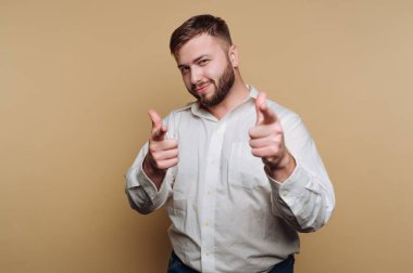 Beyaz gömlek ve kot pantolon giyen bir adam kendine güvenerek ayakta duruyor. Parmak tabancalarıyla şakacı pozlar veriyor. Cazibe ve dostluk saçıyor. Tarafsız arka plan rahat atmosfere katkı sağlıyor..