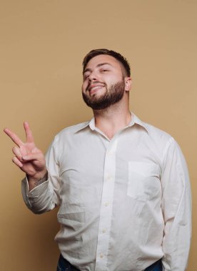 Genç bir adam dostça bir gülümsemeyle duruyor, parmaklarıyla basit bir bej arka plan karşısında barış işareti yapıyor. Beyaz gömlek ve günlük kot pantolon giyiyor..