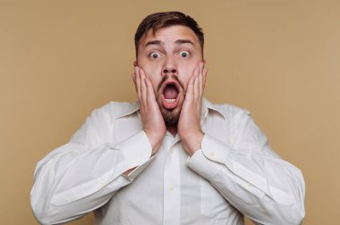 Beyaz gömlek giyen bir adam ağzı ve gözleri açık şok olmuş bir ifade sergiler. Elleri yüzüne bastırılmış, bir şaşkınlık ve merak hissi yakalamış..