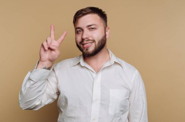 Adam büyük bir gülümsemeyle barış işareti gösteriyor. Beyaz bir gömlek giyiyor ve bej bir arka planın önünde duruyor, neşeli ve dostane bir hava yayıyor..