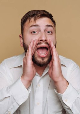 Sakallı ve kısa saçlı bir adam ellerini ağzına koyarak bağırarak heyecan ifade eder. Beyaz bir gömlek giyiyor ve yüzünde canlı bir ifade var..