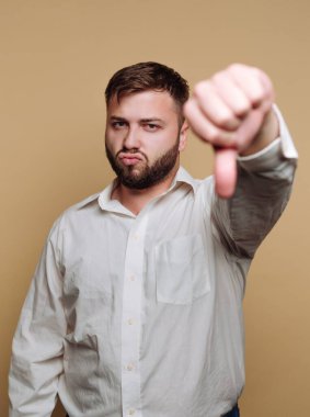 Genç bir adam beyaz gömlek giyerken baş parmağıyla bir jest yapıyor. Odaklanmış bir ifadesi var, ve arka plan basit bir bej rengi, güçlü bir görsel etki yaratıyor..