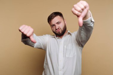 Beyaz gömlek giyen bir adam memnuniyetsizliğini baş parmağıyla bir jest yaparak gösterir. Hafifçe kaşlarını çatar, basit bir ortamda açıkça onaylamadığını belirtir..