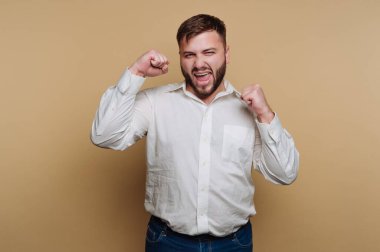 Genç bir adam sevinç ve heyecanı kutlamada yumruklarını kaldırarak ifade eder. Beyaz bir gömlek giymiş ve bej bir arka planın önünde duruyor. Pozitiflik saçıyor..