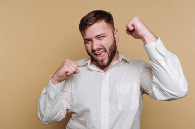 Beyaz gömlek giymiş genç bir adam, ellerini kaldırarak ve parlak bir şekilde gülümseyerek heyecan gösteriyor. Onun mutlu tavrı bir kutlama anını ya da başarısını yansıtıyor..