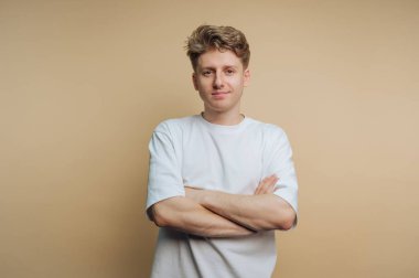 Genç bir adam kollarını kavuşturmuş, kendine güvenen bir ifade sergiliyor. Beyaz bir tişört giyiyor ve açık renkli saçları var. Yumuşak bir bej arka plana yaslanmış ve rahat duruşunu arttırıyor..