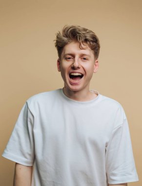 Sarı saçlı genç bir adam bol beyaz bir tişört giyerken geniş bir şekilde gülümsüyor. Tarafsız arka plan, onun neşeli ifadesini vurguluyor, dostça ve cana yakın bir hava yaratıyor..