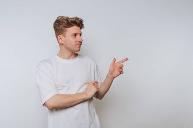 Genç bir adam güvenle ayakta duruyor, bir gülümsemeyle sağını işaret ediyor. O, basit beyaz bir tişört giydi ve sarı saç stili yaptı, rahat bir atmosfer yarattı..