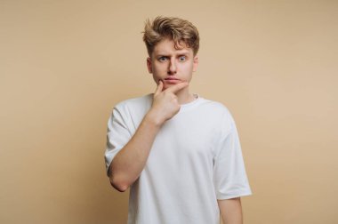 Genç bir adam tarafsız bir zemine karşı duruyor, çenesine nazikçe dokunuyor. Meraklı ve meşgul görünüyor, çözümler ya da fikirler arıyor..