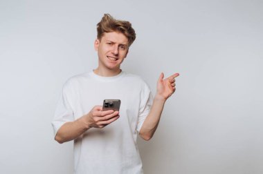 Dağınık sarı saçlı genç bir adam düz bir arkaplanda duruyor. Bir elinde akıllı bir telefon tutuyor ve diğer eliyle işaret ederken kafası karışık bir yüz ifadesi gösteriyor..