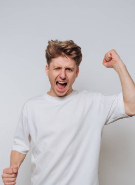 Genç bir adam yumruğunu kaldırıp bağırarak sevinç ve zafer ifade ediyor. O, sade beyaz bir gömlek giymiş, sade bir arka planı olan kapalı bir yerde. Saç stili o anın dinamik hissini arttırıyor..