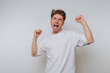 Kısa kıvırcık saçlı genç bir adam neşeli bir şekilde yumruklarını kaldırarak kutluyor. Yüzünde büyük bir gülümseme var. Beyaz bir ortamda heyecan ve coşku ifade ediyor. Muhtemelen içeride..